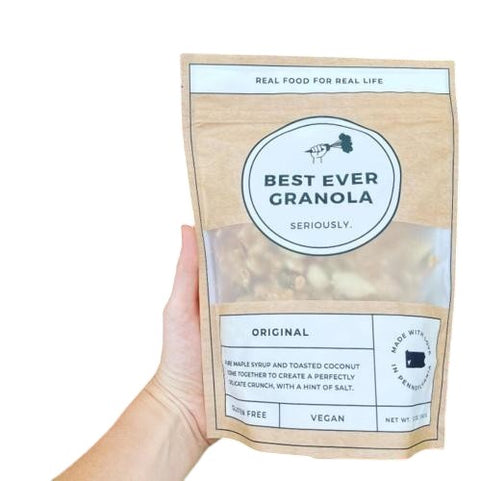 Eden’s Market Best Ever Granola dark chocolate chia flavor in a kraft paper pouch held against pink peony flowers.