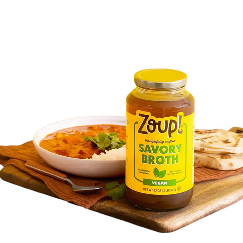 Eden’s Market Zoup! Savory Veggie Broth in a 32 oz glass jar next to a bowl of soup and flatbread on a wooden board.